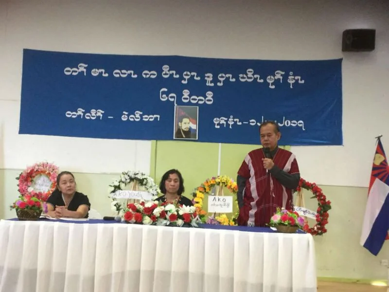 A speaker in a red Karen shirt addressing the Martyrs' Day ceremony beside a table of floral wreaths, under a banner in Karen script.