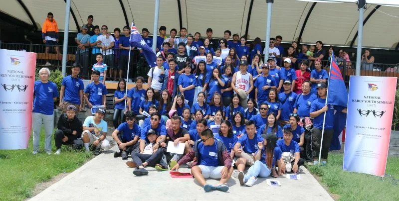 Young people in blue shirts at the 8th National Karen Youth Seminar, gathered between two seminar banners with the Australian and Karen flags.