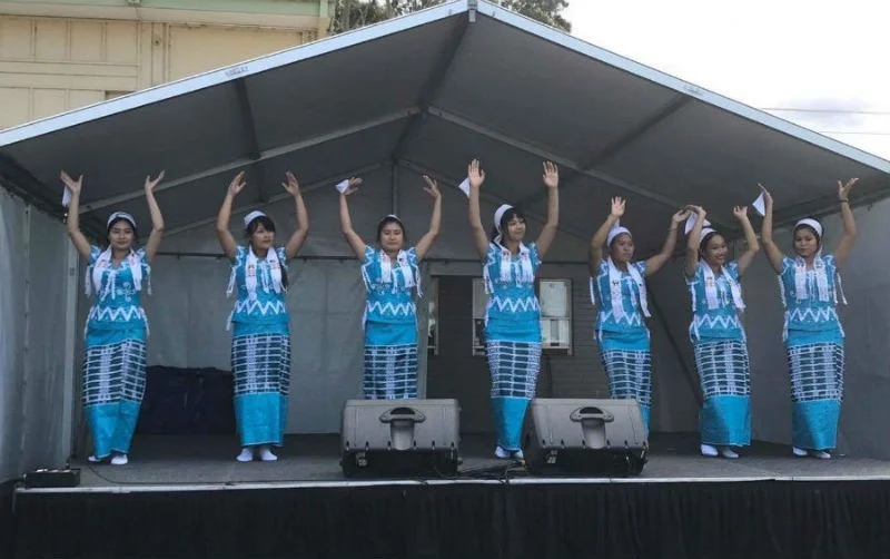 Seven women in blue traditional Karen dress dancing under a marquee at a multicultural celebration in Victoria.