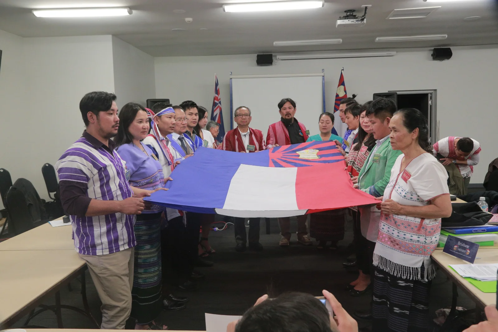 Members in traditional Karen dress holding the Karen flag outstretched in a meeting room at the 2018 National Biennial General Meeting.