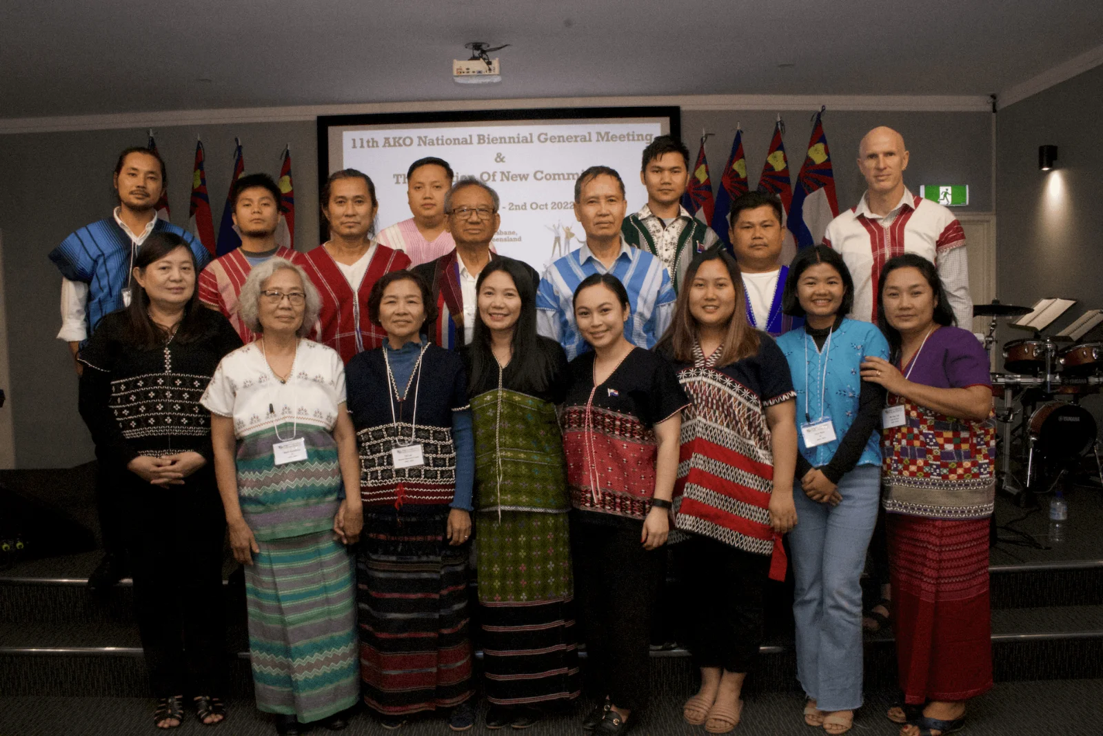 Members of the Australian Karen Organisation, many in traditional Karen dress, standing together in front of a screen announcing the 11th National Biennial General Meeting and the election of the new committee.
