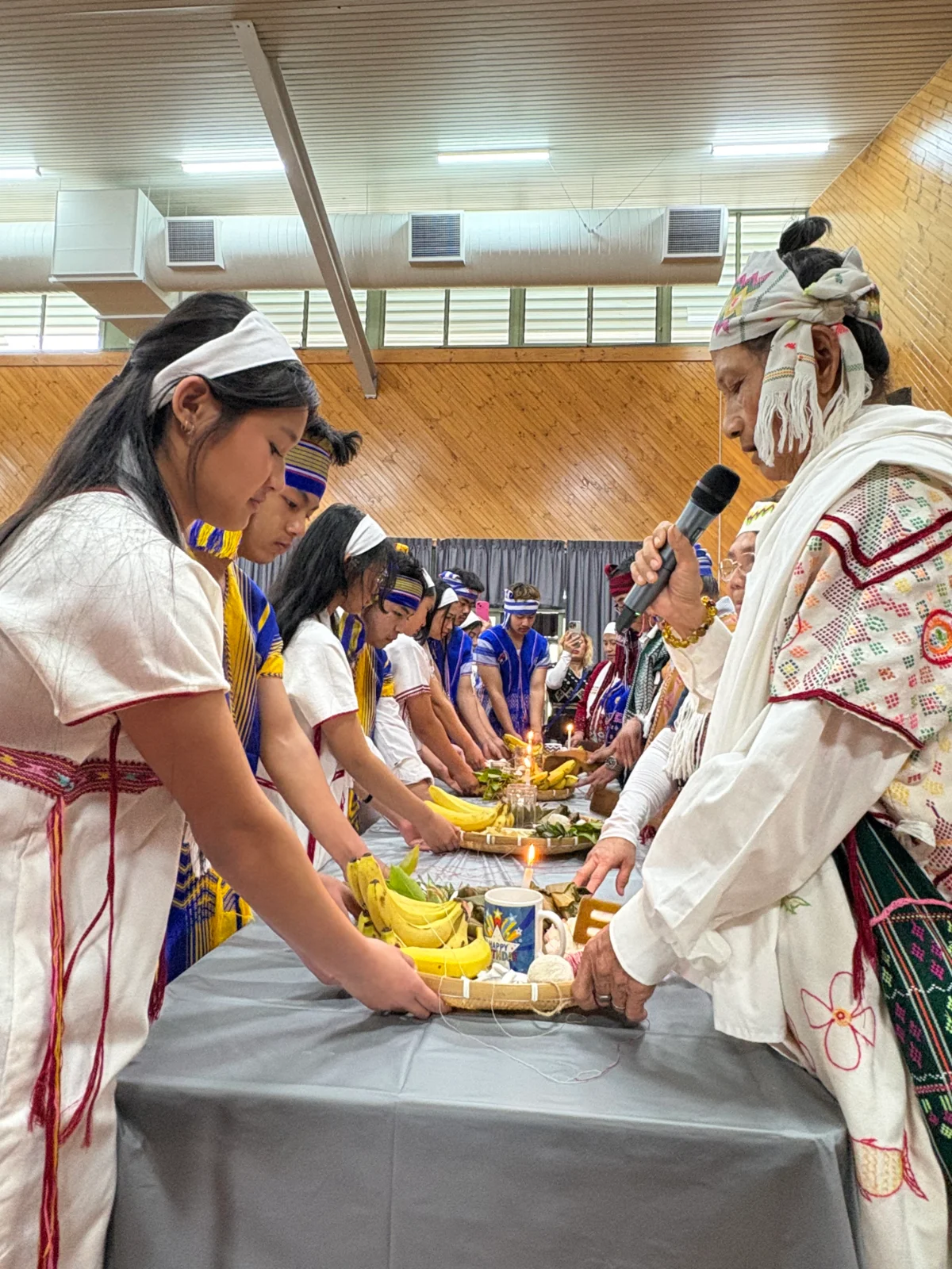 Young women in white traditional dress placing bananas and offerings on a table at the 2026 wrist-tying ceremony.