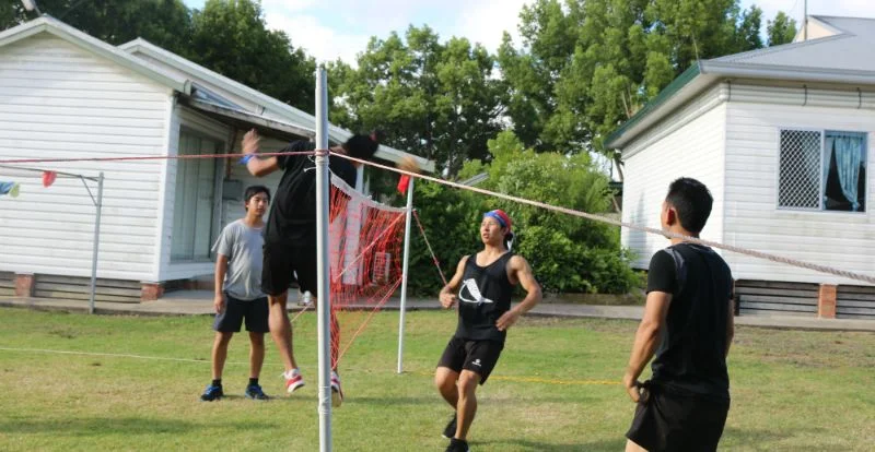 Young men playing cane ball over a net on a grassy backyard between two weatherboard buildings, one leaping for the ball.