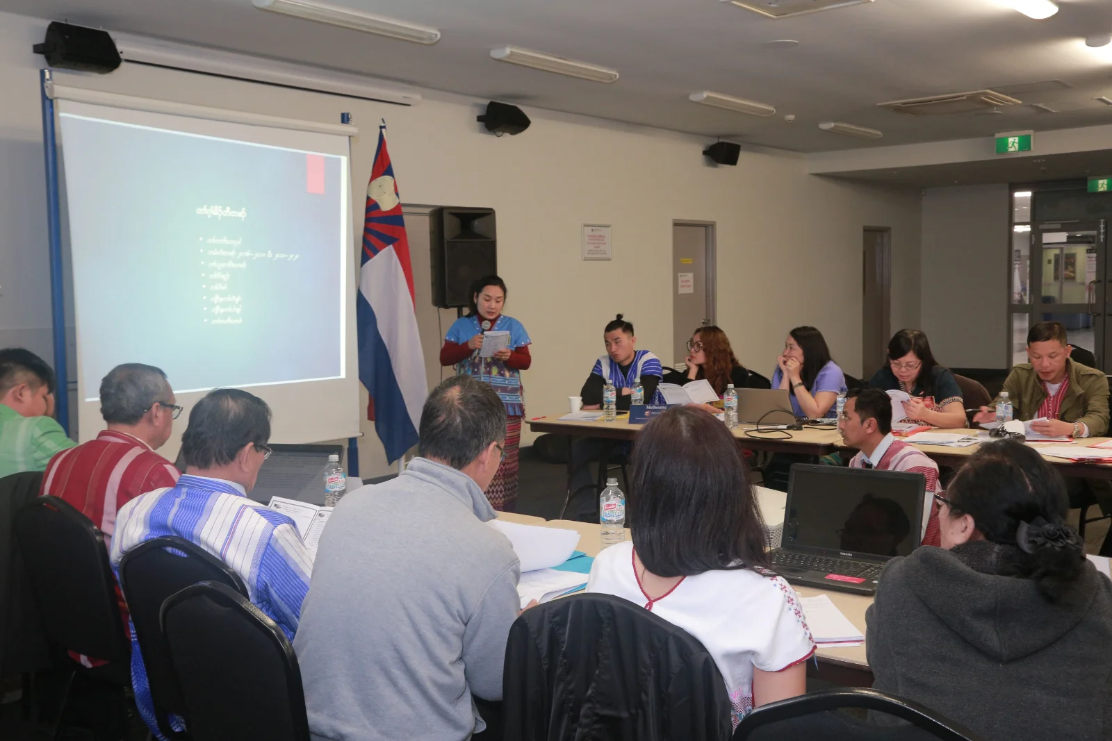 A woman in traditional Karen dress presents beside a Karen flag while committee members listen from around tables.